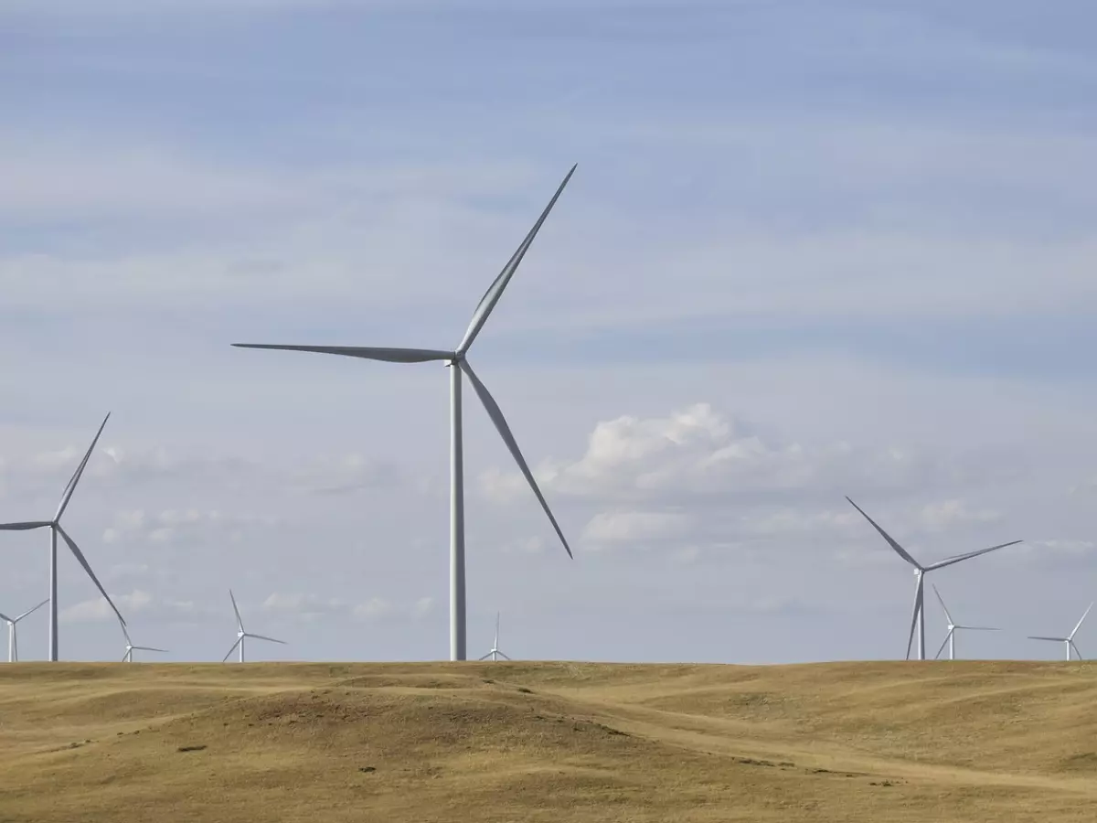 Multiple wind turbines line a vast, open field, their blades spinning against a clear, bright blue sky with fluffy white clouds. The image conveys a sense of energy and scale.